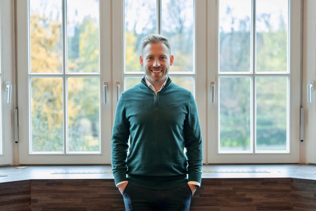 Das Bild zeigt den Gründer von Examen mit Sandro, Sandro Birkenhof. Er steht aufrecht und lachend vor einer Fensterfront mit Blick zur Kamera.